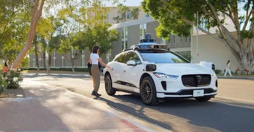 A passenger getting into a white Waymo self-driving car on a tree-lined street.
